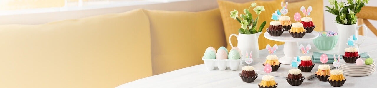 Decorated table with colored eggs, flowers and assorted flavor Bundtinis with easter eggs and bunnies toppers.
