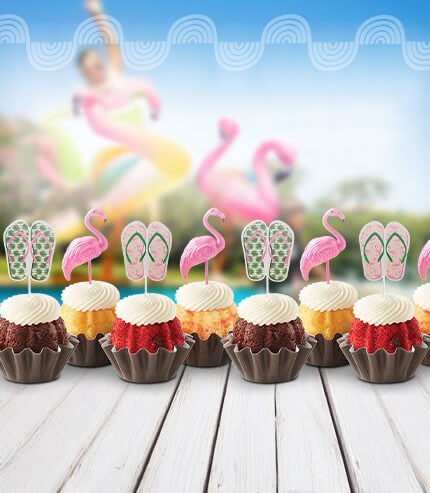 Bundtinis with flamingo and sunglasses toppers on a table poolside.