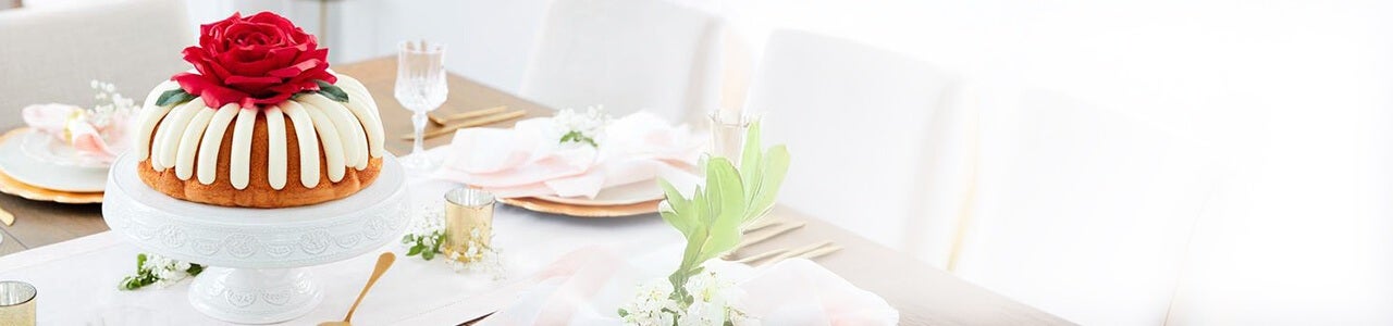 A large bundt cake with frosting, topped with a red rose decoration on a dining table.