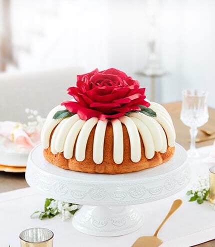 A large bundt cake with frosting, topped with a red rose decoration on a dining table.