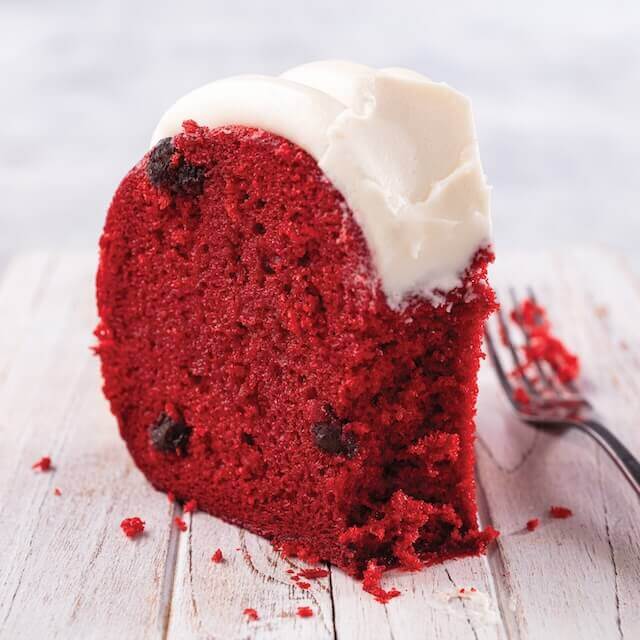 Red Velvet Bundt cake crowned with signature cream cheese frosting on a cake stand and a slice taken out showing chocolate chips baked into the cake - Shop Bundt Cakes