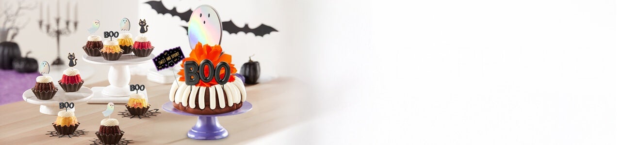 Assorted bundtinis and a bundt cake with holographic ghosts, black cat cutouts and boo sign decorations.