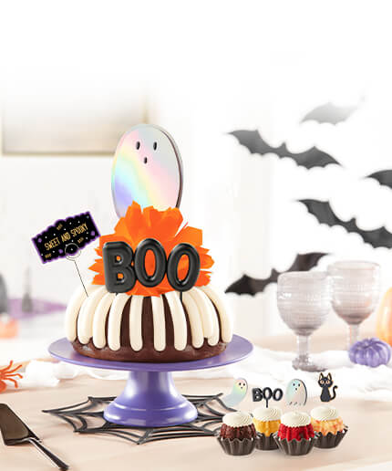 Assorted bundtinis and a bundt cake with holographic ghosts, black cat cutouts and boo sign decorations.