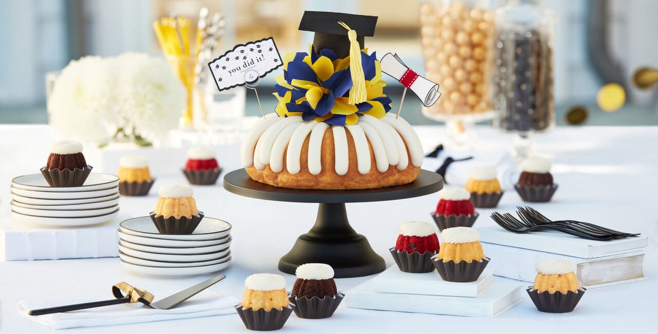 Graduation celebration dessert table featuring Bundt Cake with cream cheese frosting, graduation cap, blue and yellow tissue paper flower, diploma decoration, and you did it sign, surrounded by assorted Bundtinis with cream cheese frosting for graduation parties and special occasions - Shop Now