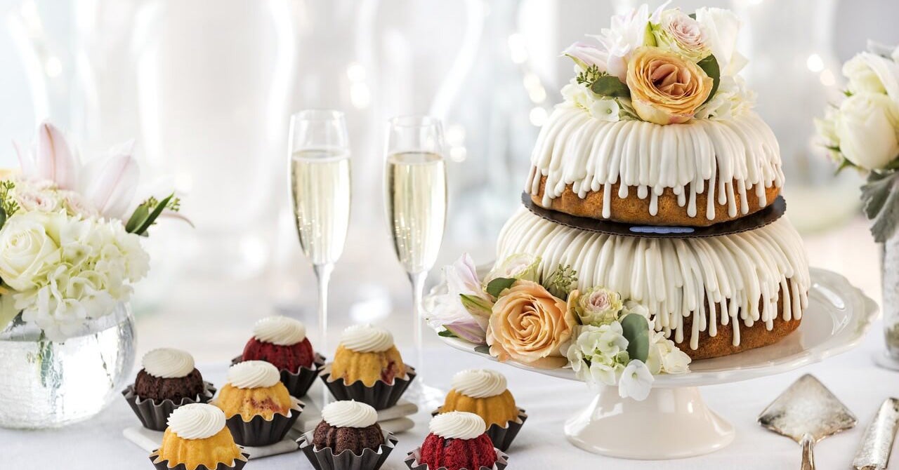 Elegant wedding dessert display featuring tiered Bundt Cakes with cream cheese frosting and fresh roses, surrounded by assorted Bundtlets with cream cheese frosting, champagne glasses, and white floral arrangements for wedding celebrations. Shop Now.