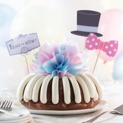 A gender reveal bundt cake with a decoration of a top hat and a pink dotted bow tie. A card reads Beau or Bow? and the center of the cake has a pink and blue bow.