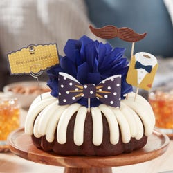 A bundt cake with frosting and topped with a bowtie and a moustache decoration. A card is attached to the cake and reads best dad a'round.