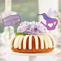 Large cake with frosting on top. The center of the bundt cake is covered with a silk hydrangea flower. There is a hummingbird cutout also decorating the cake and a card that reads best mom a'round.