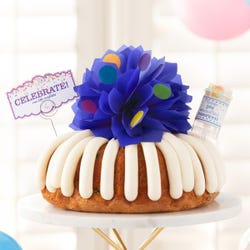 Large decorated bundt cake covered with frosting. Decoration includes a pink bow with confetti sprinkles and a confetti popper with a card that says Celebrate and cue the confetti.