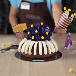 A bundt cake with a navy colored bow with golden pom-poms. A nay and yellow crown tops the cake along with card that says c'round' for the day!.