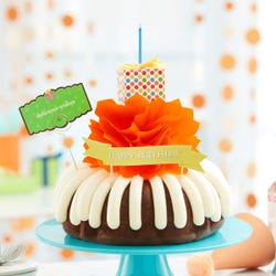 A birthday cake with frosting on top. The cake is decorated with an orange bow and a wrapped gift box with different color dots and topped with a gift bow and a candle.