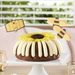 A large bundt cake with frosting. The center of the cake is topped with a large sunflower and adorned with bumblebee cutouts and a card that reads flour bouquet - Thinking of you.
