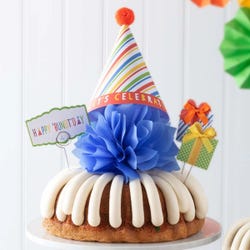 A bundt cake with frosting topped with a party hat that says Let's Celebrate. Two wrapped gift boxes are on the sides along with a card that reads Happy 'Bundt'day.