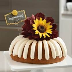 A bundt cake with frosting with a chocolate/brown colored bow and a sunflower topper. There is a decoration card that says happy everything! with a smiley face.