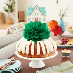 Bundt cake topped with a green pom bow and a small house decoration above it. There are two mini balloons decorating the home along with a card that reads Home Sweet Home.