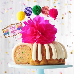 A party-themed bundt cake with colorful 3D balloons. The cake has a card that reads LET'S PARTY.