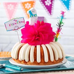 A party-themed bundt cake with a colorful mini piñata at the center and a piñata stick next to it. The cake has a card that reads FIESTA. 
