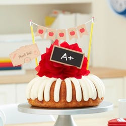 A frosted bundt cake with a red bow in the center. The cake is decorated with a chalkboard with an A+ written on it along with a card that reads Thank You!.
