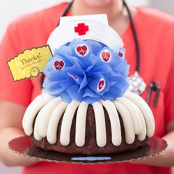 A cake with frosting and topped with a blue bow with heartbeat stickers and a nurse hat at the top. A card reads Thanks! You take the cake.