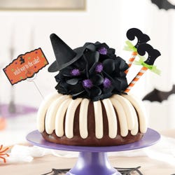 Last Chance! This bundt cake is decorated with frosting all around. It is topped with a black bow with a witch hat and legs sticking out of the cake. A card is attached and reads witch way to the cake?.