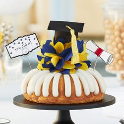 Bundt cake with frosting and topped with a customizable colored bow. It is topped with a diploma rolled up and a graduation cap with a tassel and a card that says you did it!.