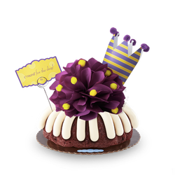 A bundt cake with a plum colored bow with golden pom-poms. A plum and yellow crown tops the cake along with card that says c'round' for the day!.