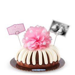 A bundt cake with a pink bow in the center with a small photo card of baby shoes and a pink dotted card that reads baby cakes. Sweet congratulations!