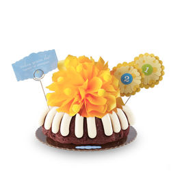 Birthday bundt cake with frosting all around. Topped with a golden yellow bow and numbered flowers suitable for age or any numbered milestone. 